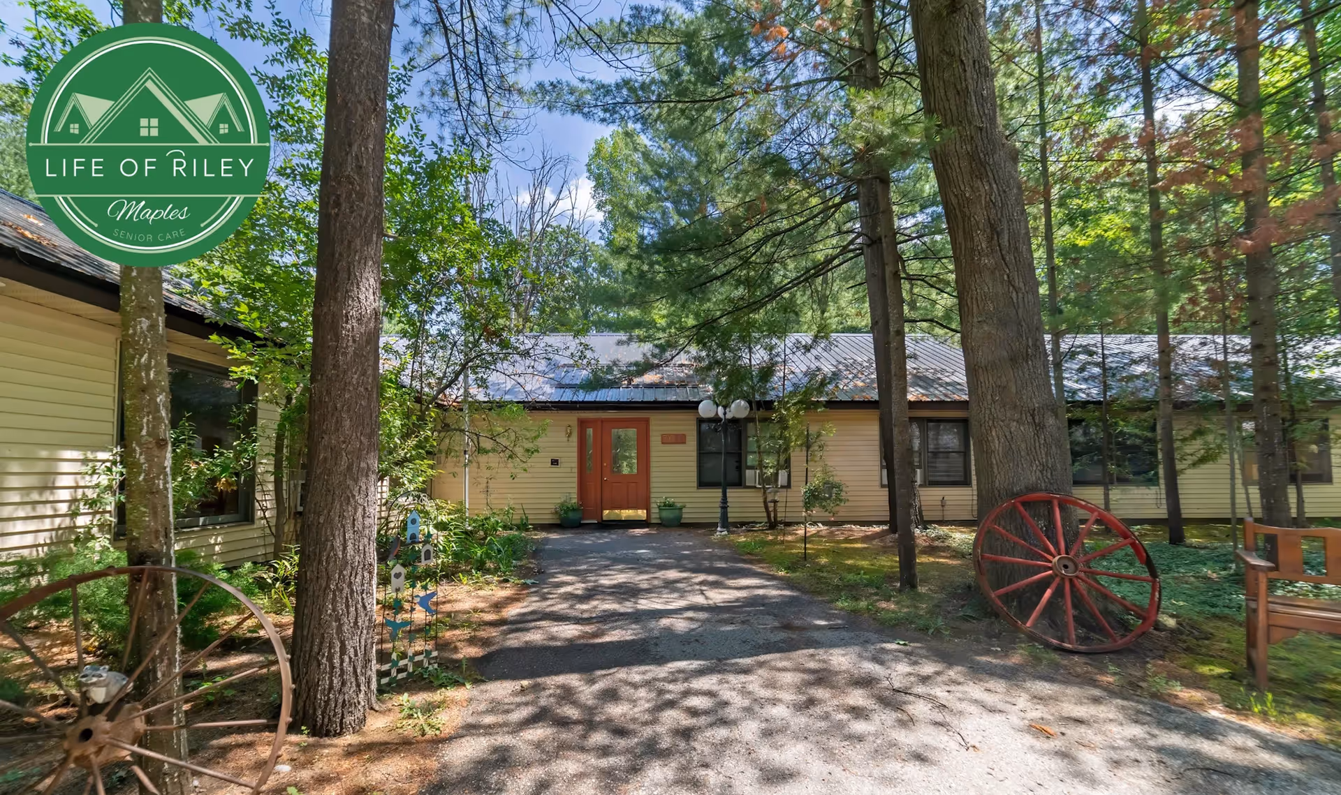 Front exterior of a single-story senior care building with trees and outdoor seating.