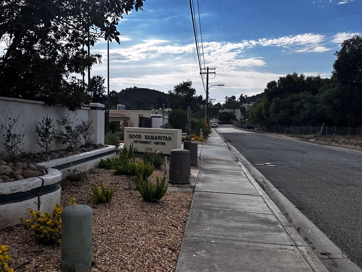 Sidewalk and street view showing the front sign for Good Samaritan Retirement Center with landscaping and utility poles under a partly cloudy sky.