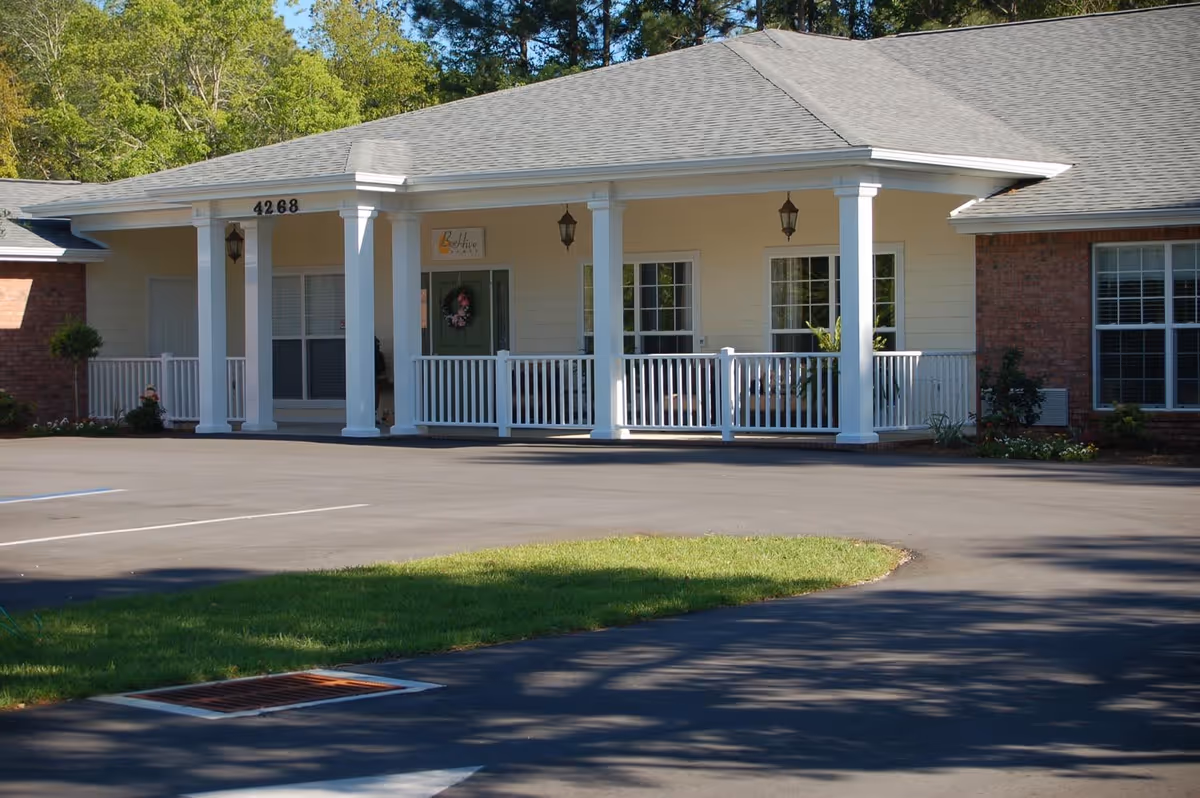 Front exterior of a single-story assisted living building with a covered porch, white columns, railing, and a paved parking area.