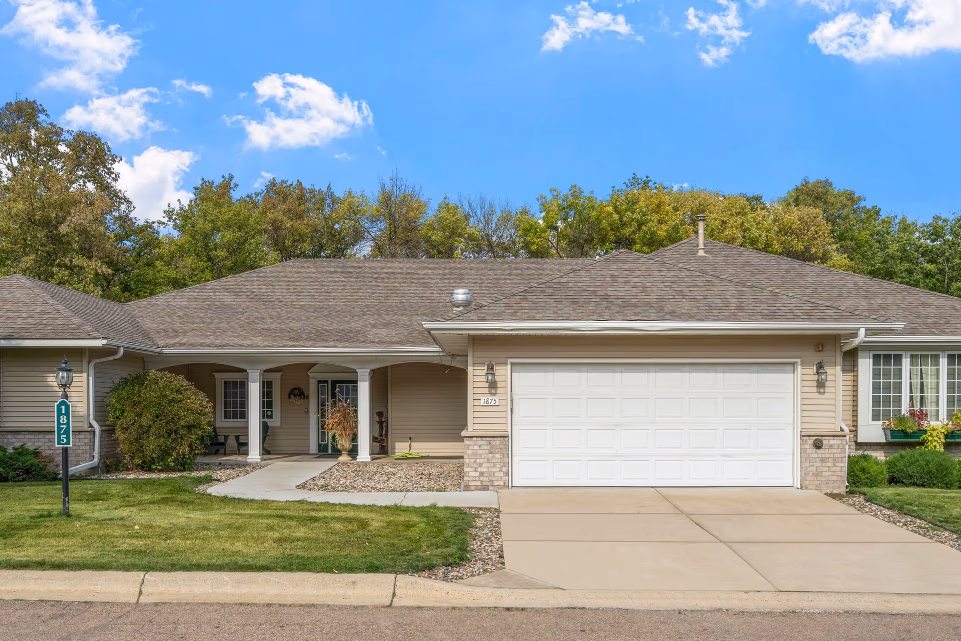 Exterior front view of a single-story residential-style building with a two-car garage, a covered porch with white columns, and a well-maintained lawn and landscaping under a blue sky with some clouds.