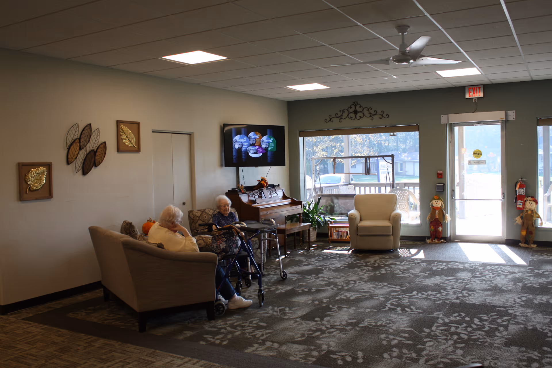 Senior residents sit on couches in a spacious lounge with a TV and piano.