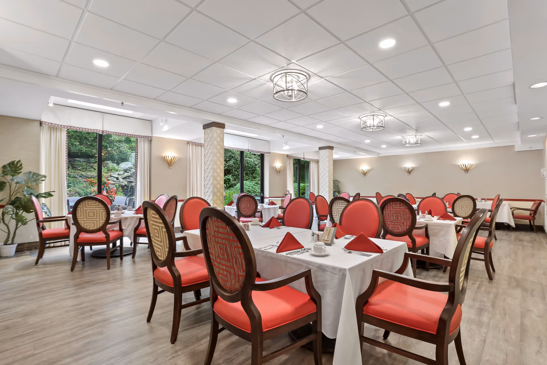 A bright and elegant dining room with multiple tables covered in white tablecloths, each set with red napkins, cups, and silverware. The chairs have wooden frames with red and patterned upholstery. Large windows reveal a green outdoor garden area, and the ceiling features modern light fixtures. The room has a warm and inviting atmosphere.