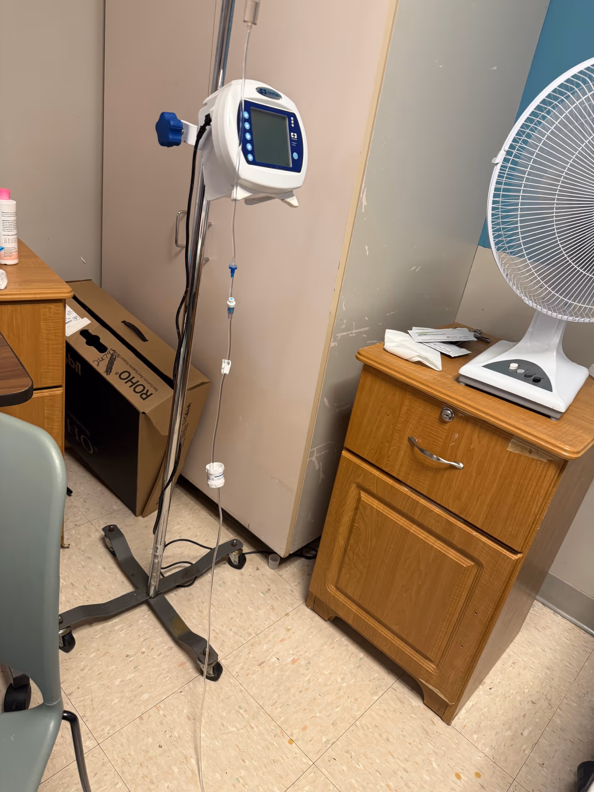 IV infusion pump on a pole beside a cabinet and a fan in a clinical room.