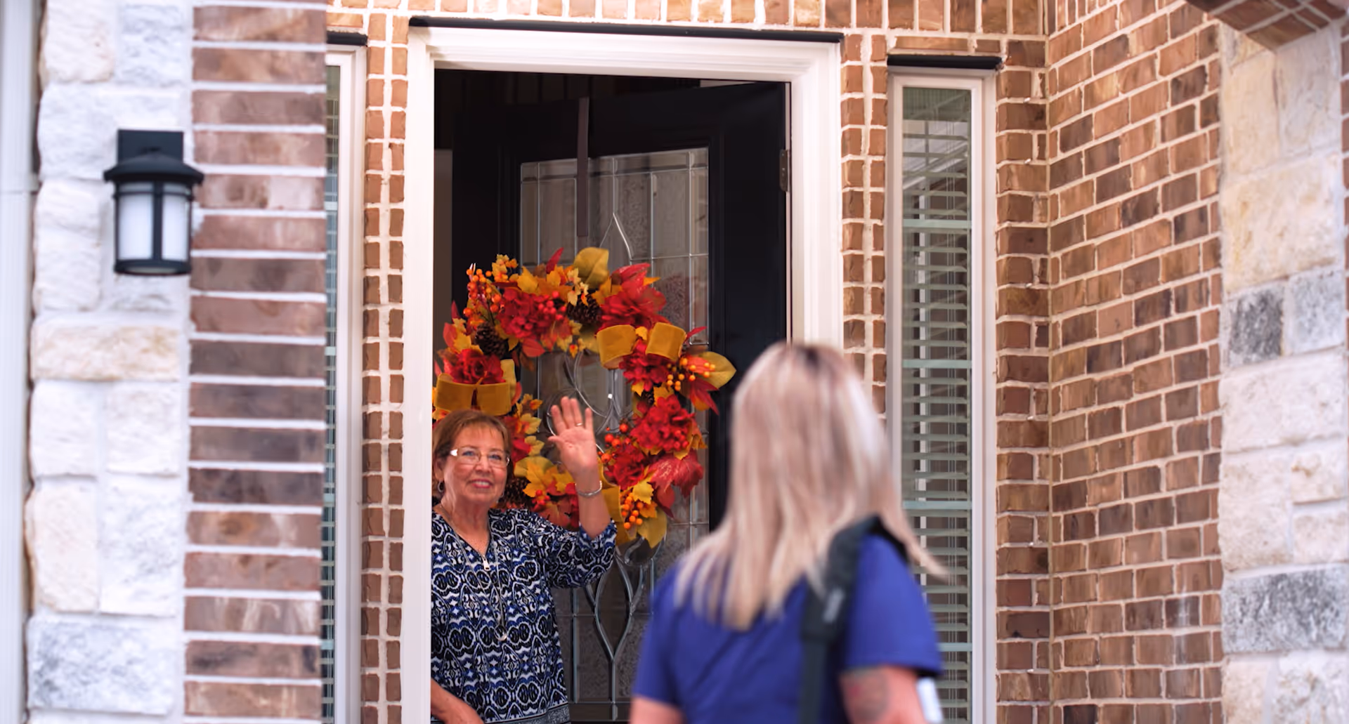 A woman waves from a brick front door entrance with a colorful autumn wreath.