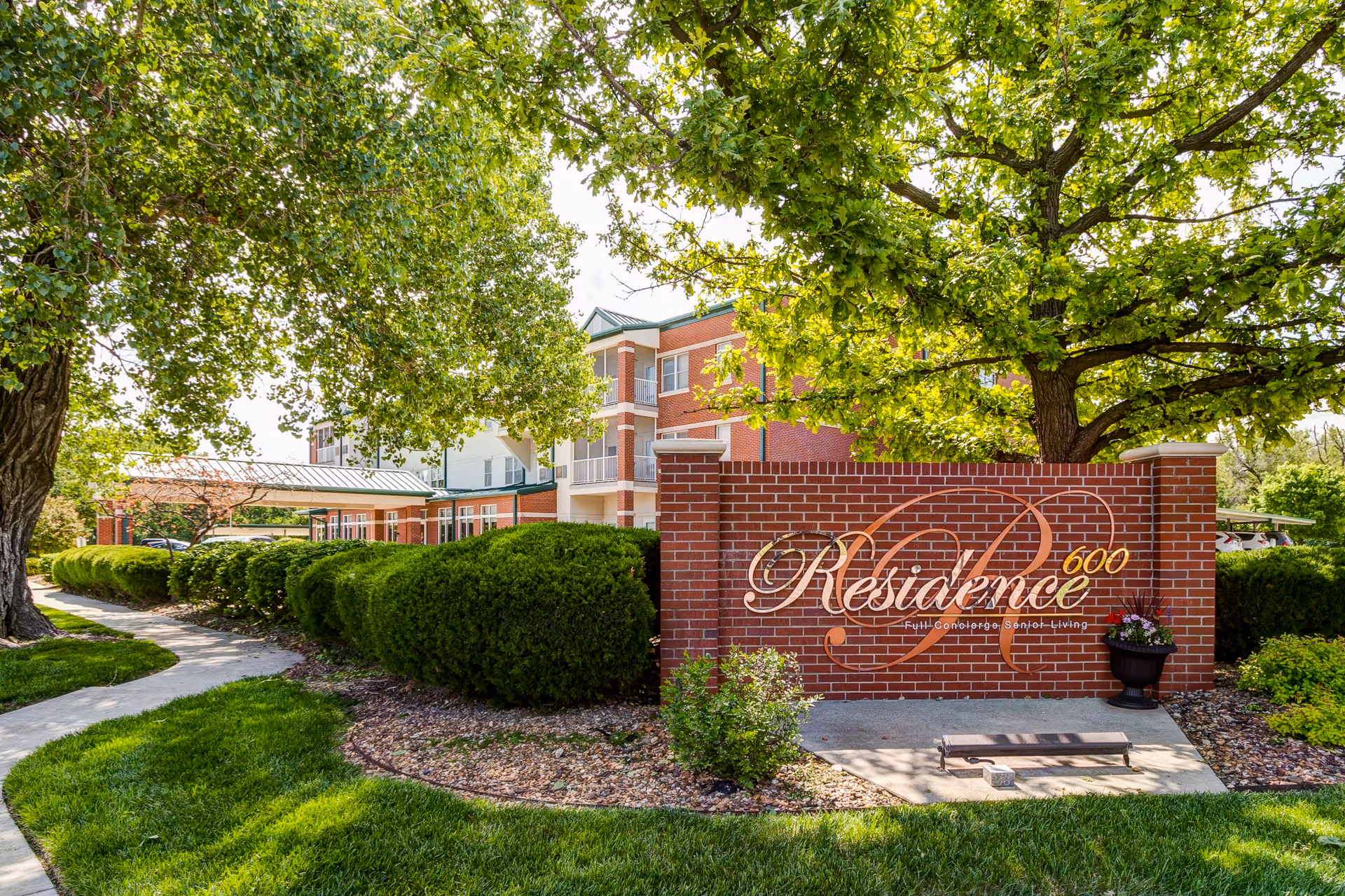 Exterior view of Residence 600 senior living facility showing a brick sign with the facility name, surrounded by green bushes and trees, with a sidewalk curving through a well-maintained lawn.