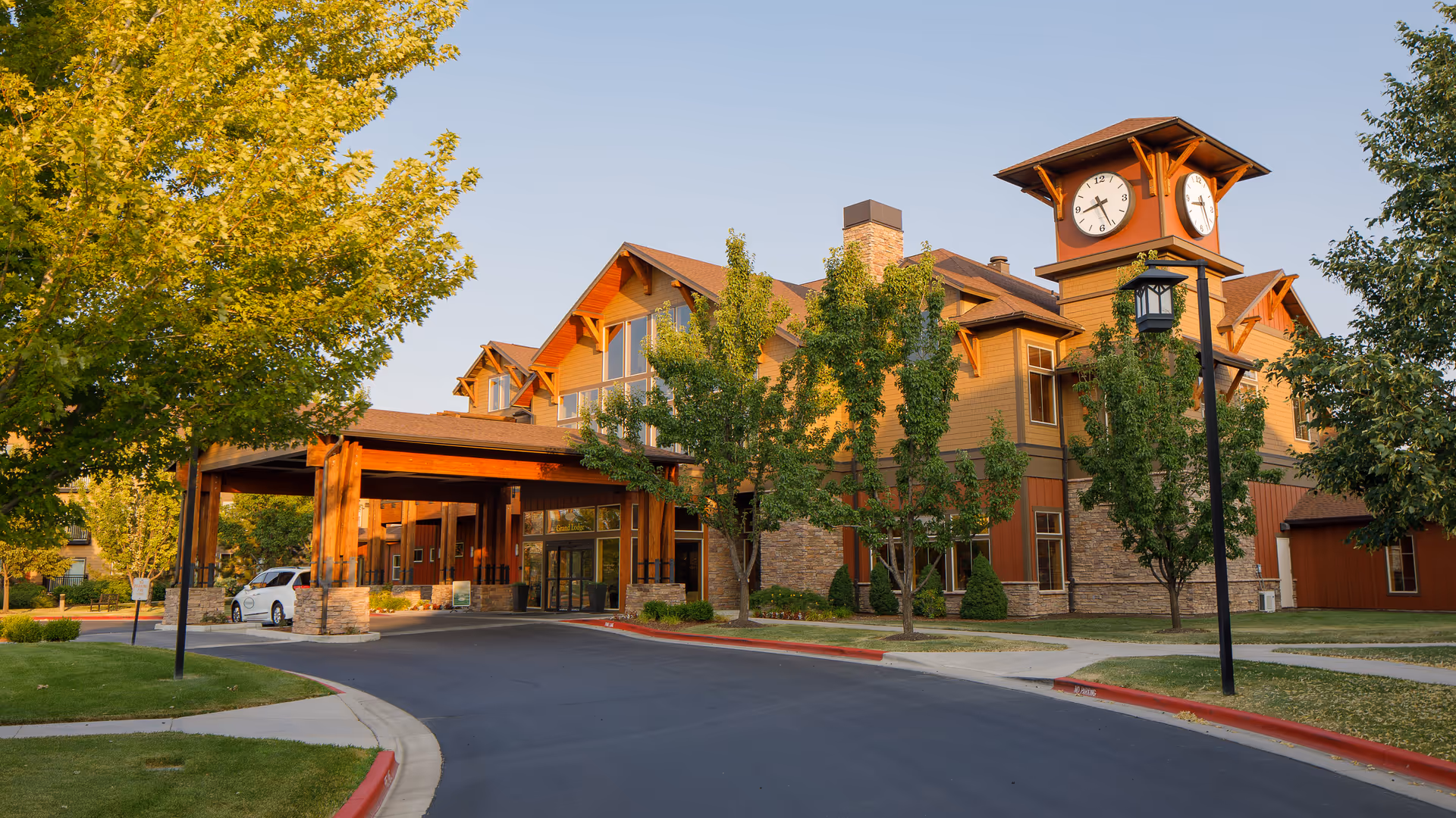 Front exterior of a multi-story community building with a covered entrance, clock tower, trees, and a driveway.