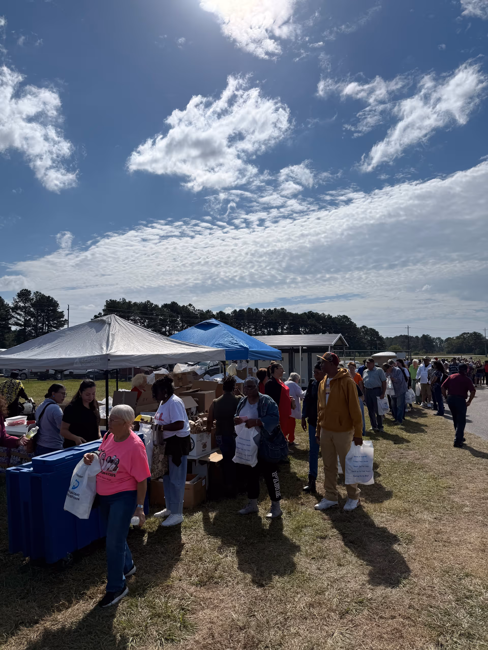 Outdoor community event with tents, volunteers, and people lining up for donations.
