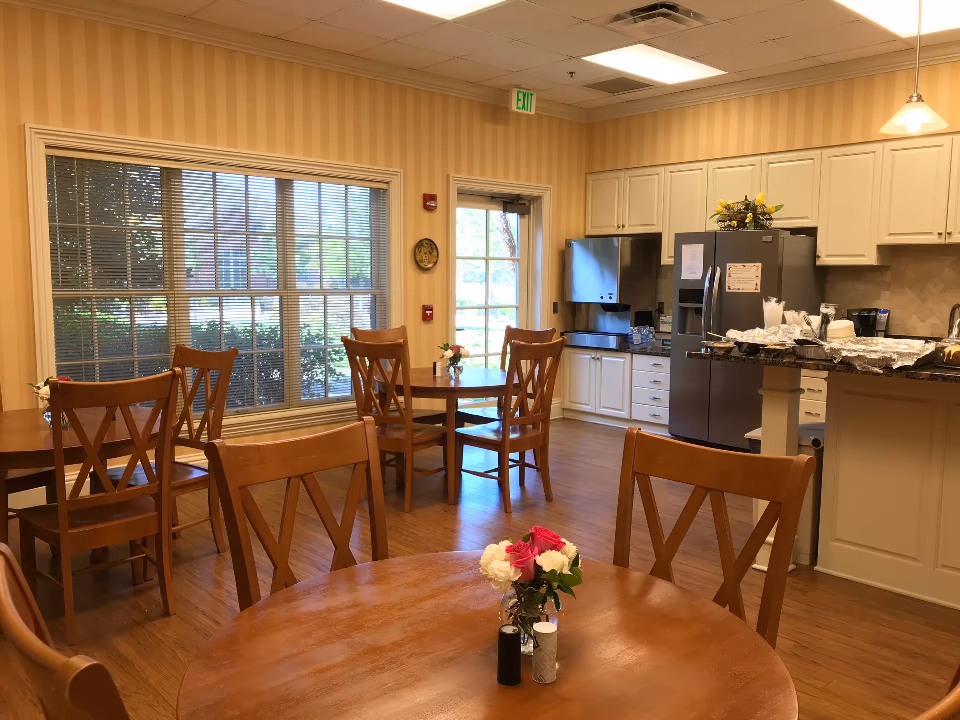 Bright dining area with round wooden tables, chairs, and a connected kitchen in a senior facility.