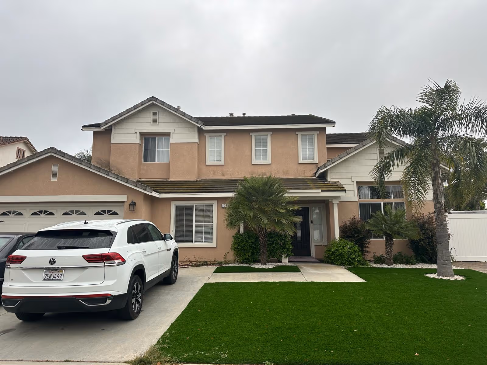 Two-story beige house with a driveway, palm trees, and a white SUV in front.