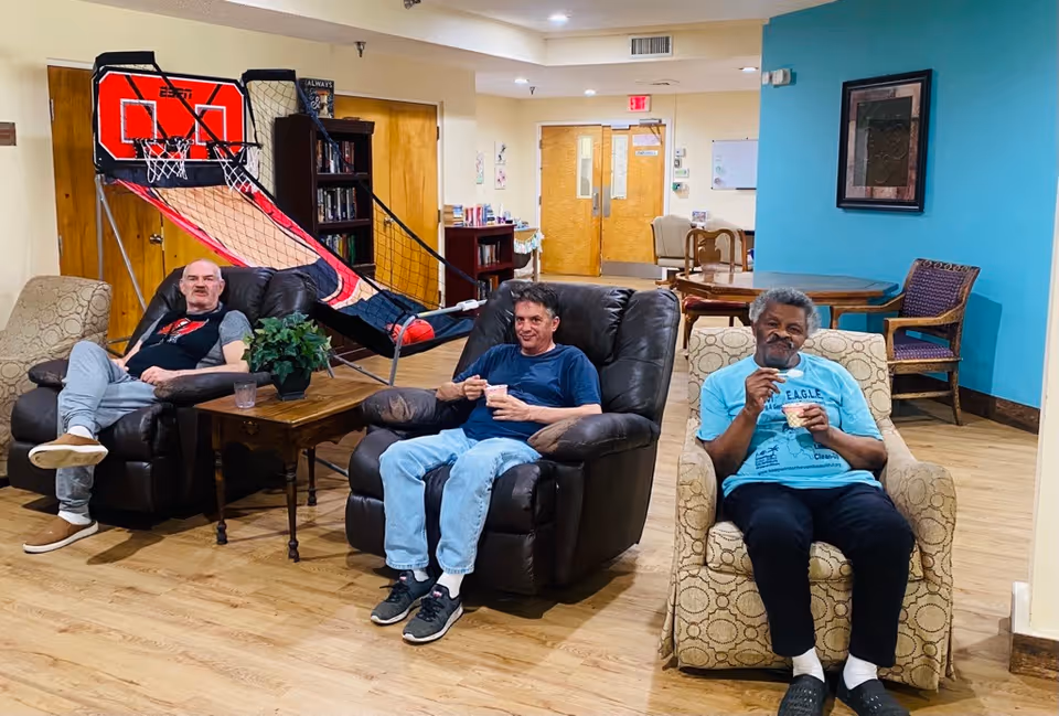 Three elderly men sitting comfortably in armchairs in a common area of a senior living facility. Behind them is a basketball arcade game, bookshelves, and a table with chairs. The room has wooden flooring and light-colored walls with a blue accent wall.