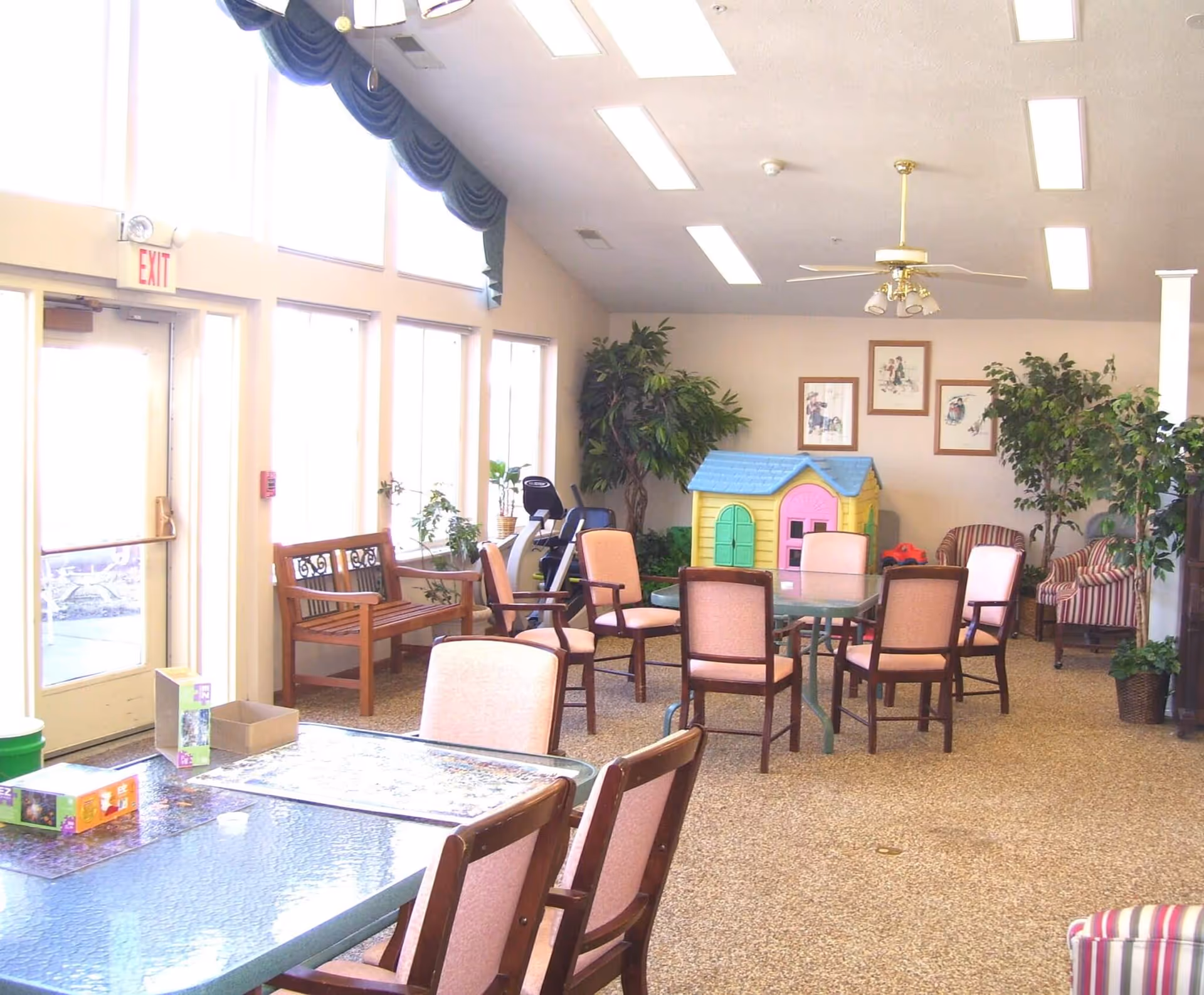 Bright communal activity room with tables and chairs, potted plants, a small colorful playhouse, and large windows along one wall.