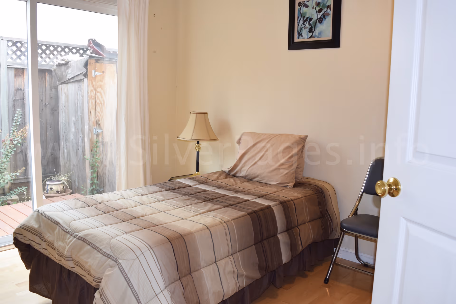A small bedroom with a single bed, lamp, and chair beside a sliding door.