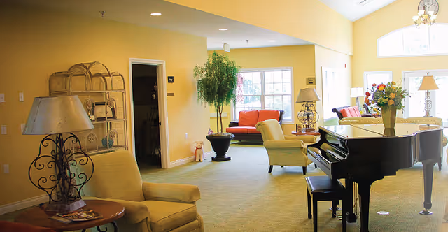 Bright communal lounge with upholstered chairs, a grand piano, side tables and lamps near large windows.