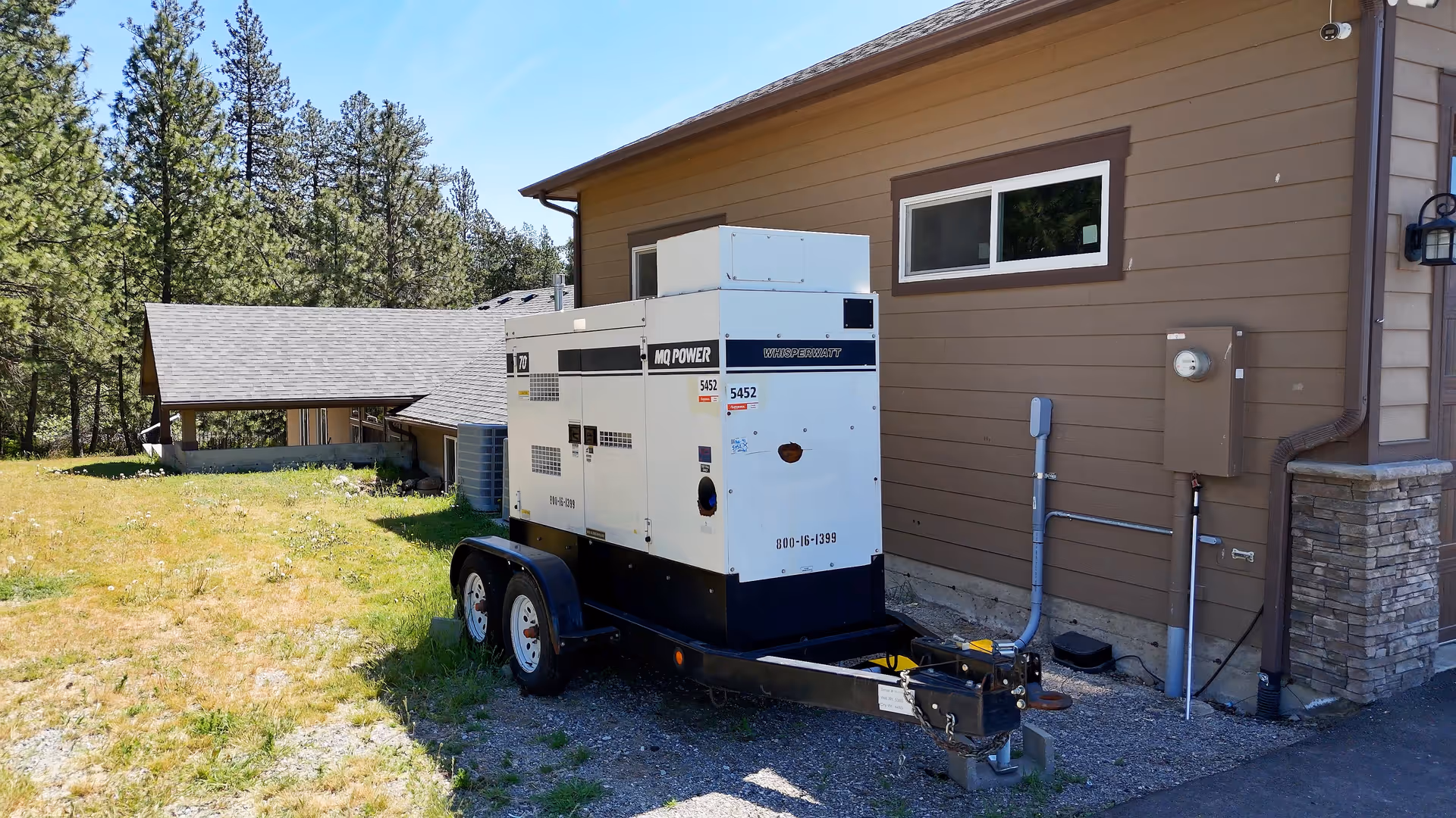 A white generator on a trailer parked beside a brown building outdoors.
