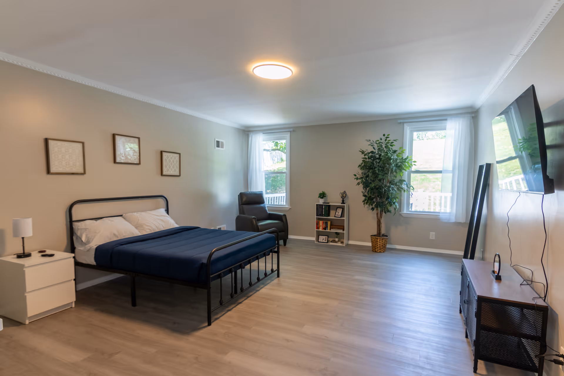 Neutral bedroom with metal bed, nightstand, armchair, bookshelf, plant, and wall-mounted TV.