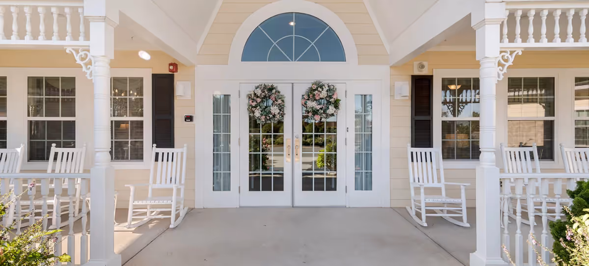Front entrance of a senior living facility with double glass doors decorated with floral wreaths. The entrance is flanked by white rocking chairs on a covered porch with white railings and columns. Windows with black shutters are visible on either side of the doors.