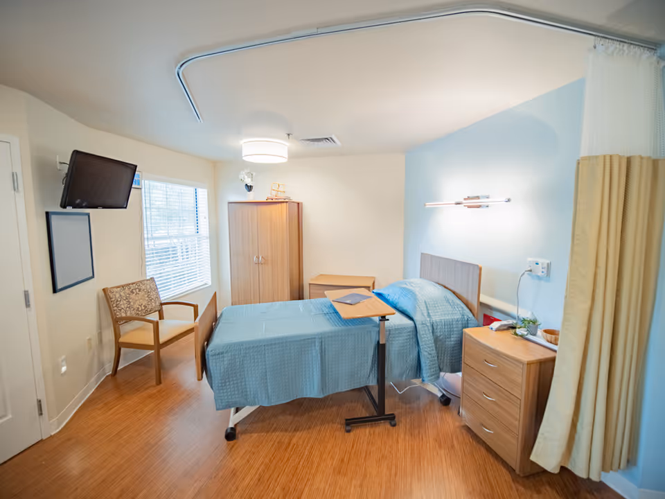 Hospital-style patient room with a blue bed, overbed table, wardrobe, chair, and bedside dresser.