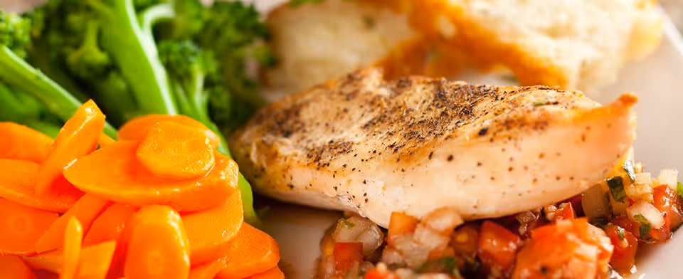 Close-up of a plated meal featuring a seasoned grilled chicken breast served with cooked sliced carrots, steamed broccoli, and a side of diced tomato and onion salsa.