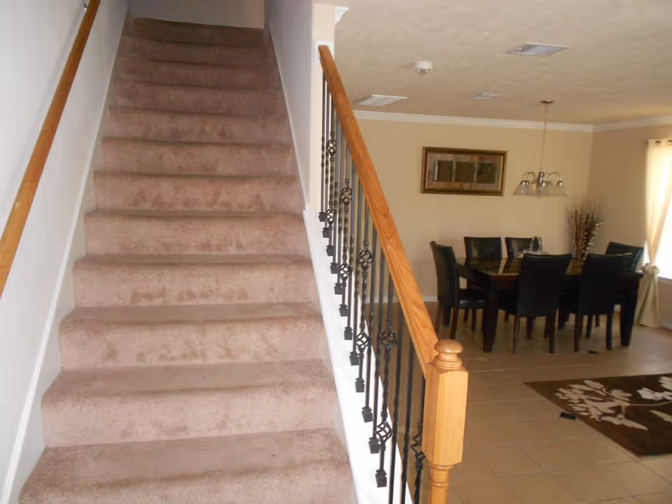 Carpeted staircase with a wooden banister and wrought-iron spindles leading up, adjacent to a dining area with a table and chairs.