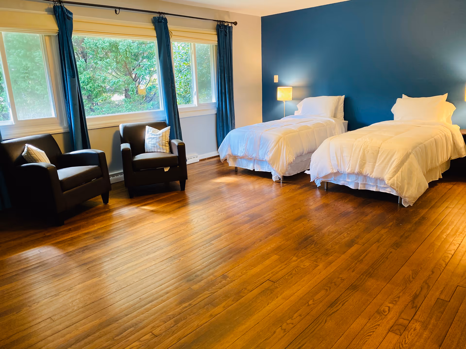 Two beds with white comforters, blue accent wall, and chairs by a large window.