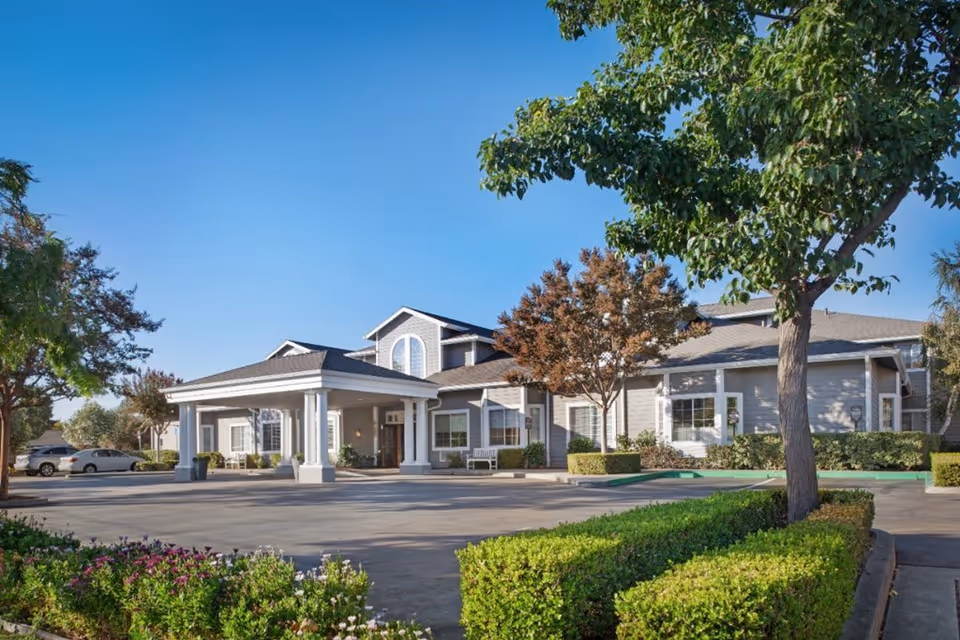Exterior view of a senior living facility building with a covered entrance, surrounded by trees, shrubs, and a clear blue sky.