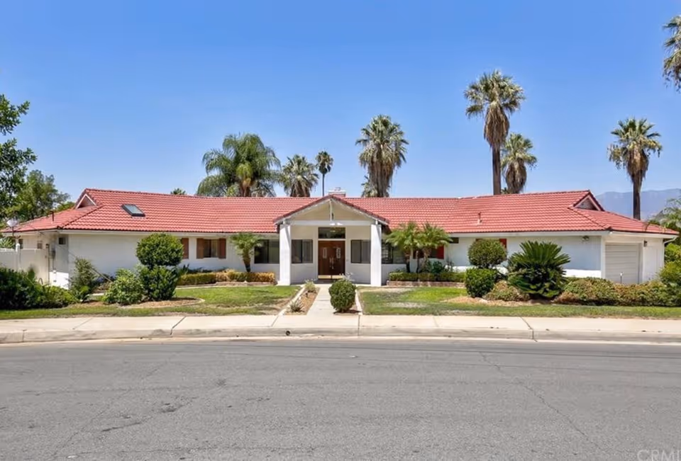 Single-story white building with a red tile roof, landscaped front yard and palm trees under a clear blue sky.