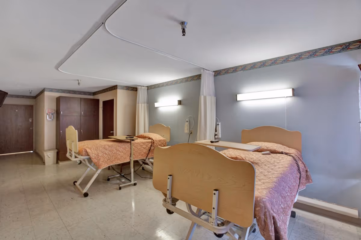 A room with two hospital-style beds, each with a peach-colored quilted blanket, adjustable overbed tables, and wall-mounted lights above the headboards. The room has a tiled floor, a wooden cabinet, and privacy curtains between the beds.