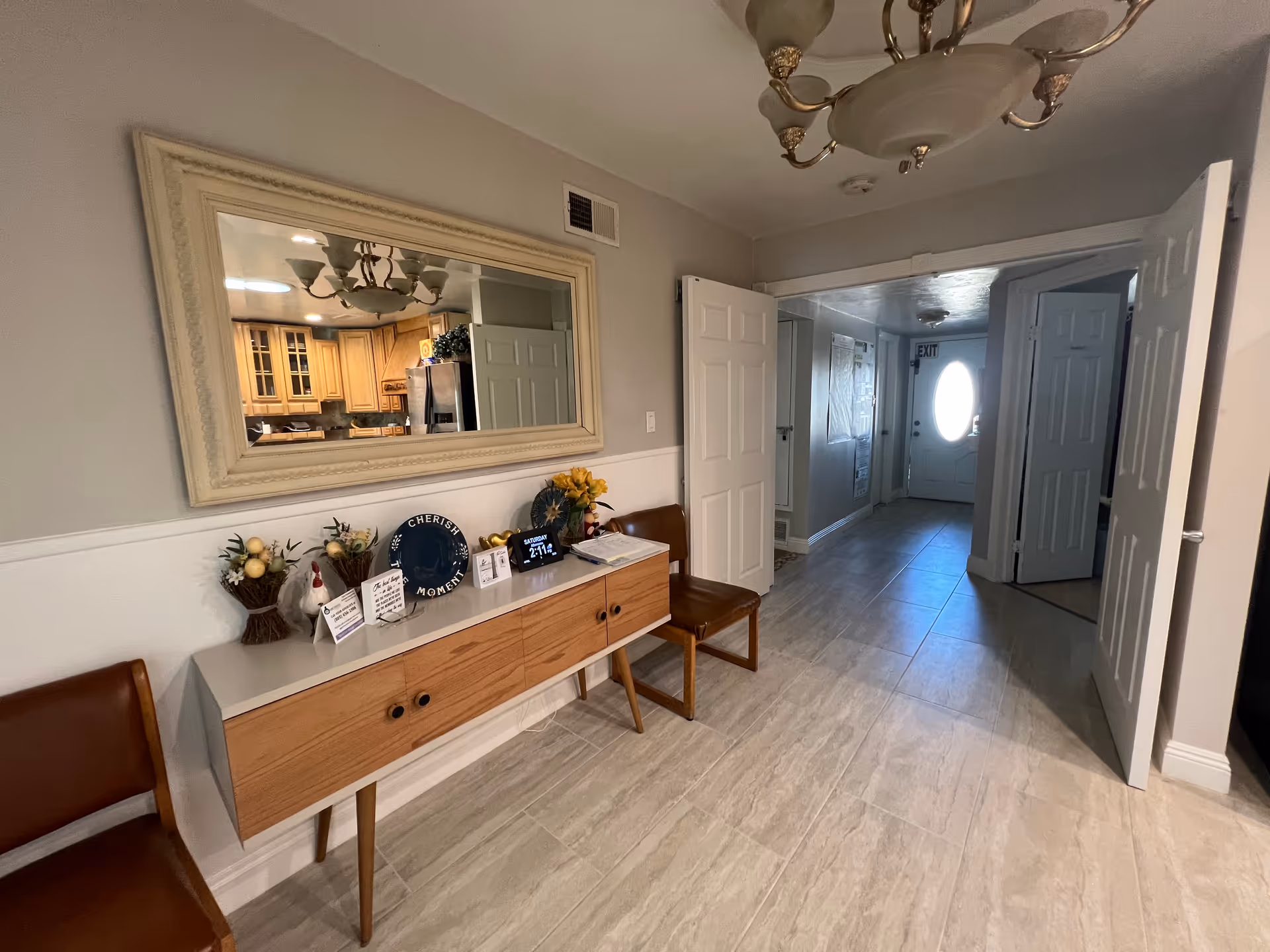 Interior hallway area with light-colored tiled floor, a wooden console table with decorative items including flowers, a plate, and framed notes, two brown chairs on either side of the table, a large rectangular mirror above the table reflecting a kitchen area, and an open doorway leading to a hallway with a white door featuring an oval window and an exit sign above it.
