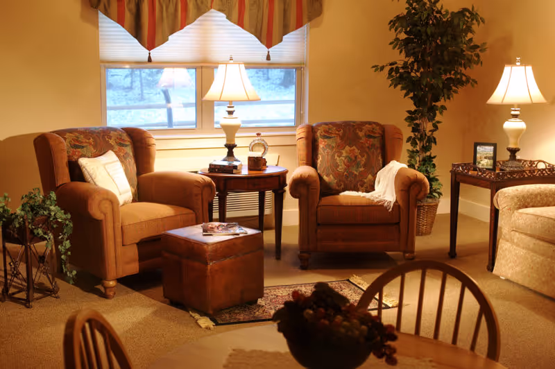 Cozy living room area with two upholstered armchairs, a small round wooden table with a lamp and decorative items between them, a leather ottoman, a potted plant, and a side table with another lamp and framed photo. A window with blinds and valance is in the background, and part of a dining table with chairs is visible in the foreground.