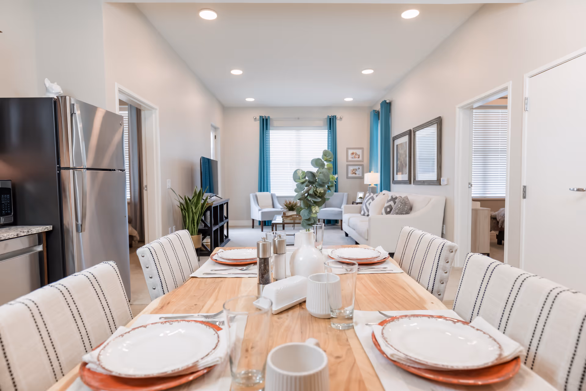 Bright open-plan dining area with a set wooden table in the foreground and a living room with sofa and TV in the background.