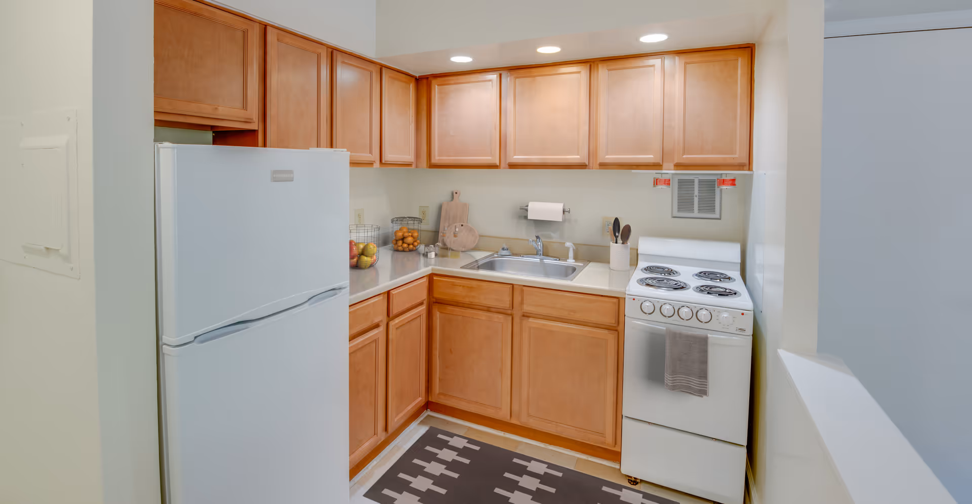 Compact kitchen in Victory Forest senior living facility with wooden cabinets, refrigerator, sink, and stove.