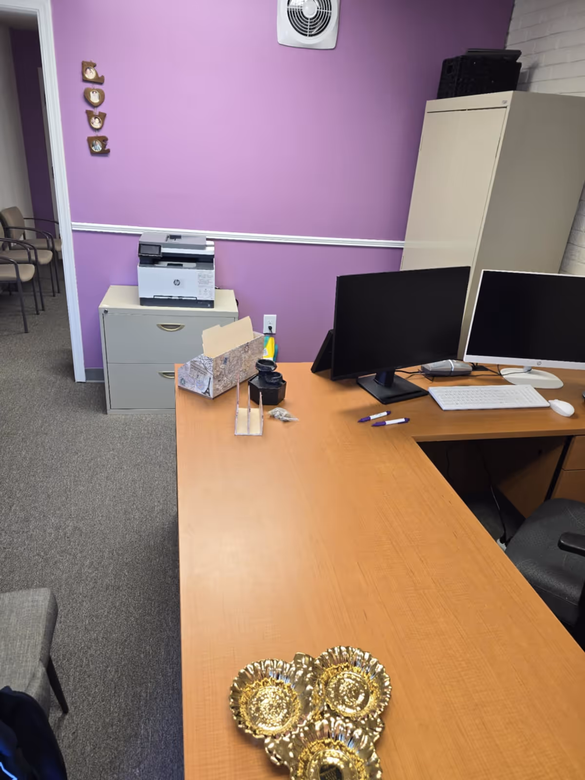 Office desk with two monitors, printer, purple wall, and filing cabinet in a care facility.