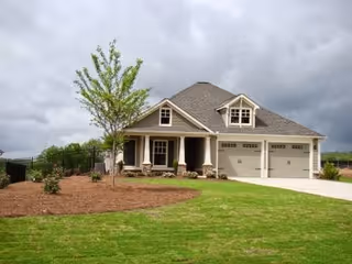A single-story house with a gray roof, beige exterior walls, and a two-car garage. The house has a front porch with columns and a small tree planted in the front yard. The sky is cloudy and the lawn is green and well-maintained.