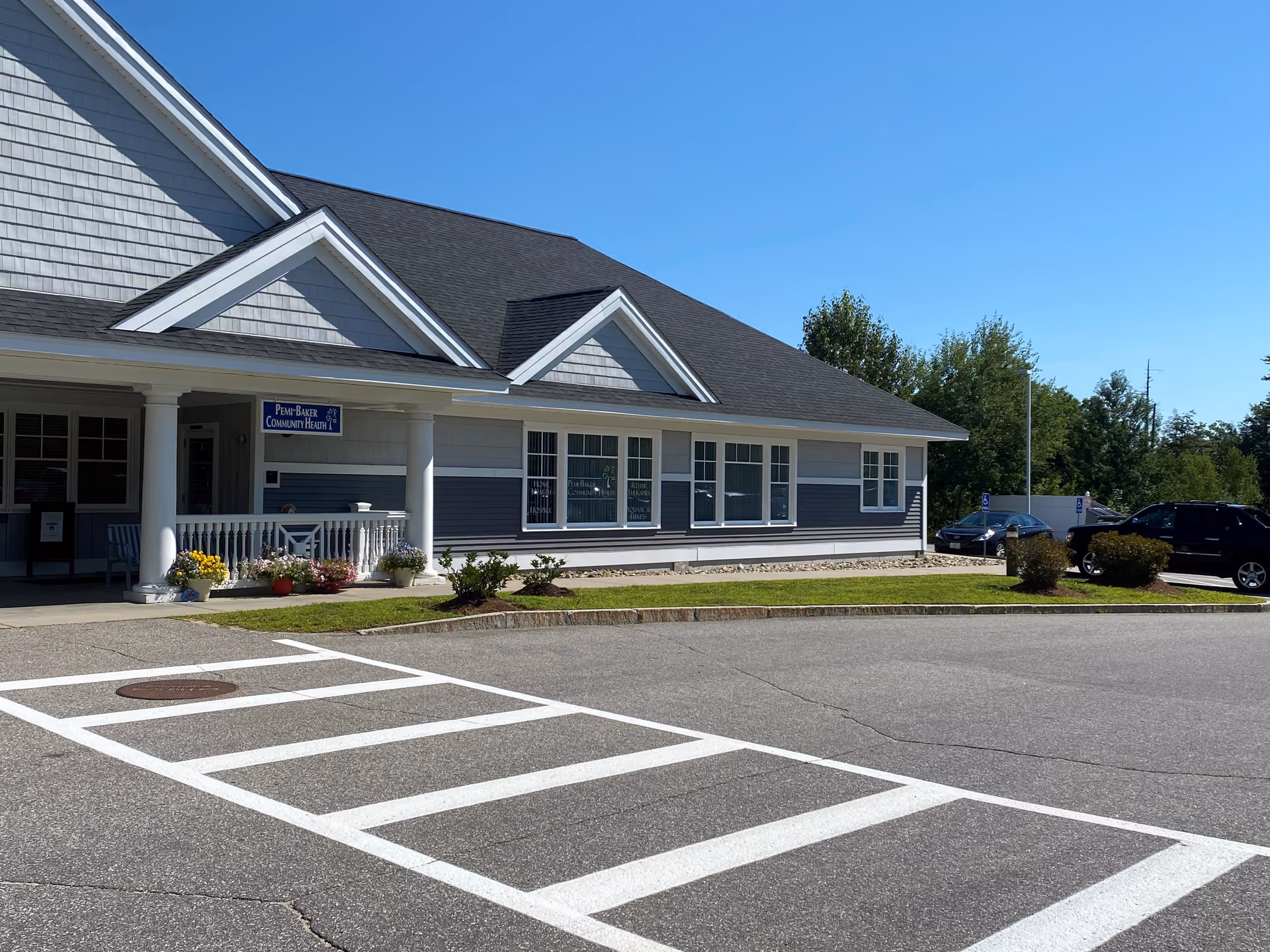Blue-gray hospice building with white trim, entry porch, and parking lot under a clear sky.