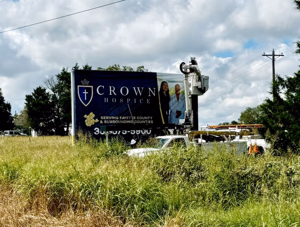 An outdoor Crown Hospice billboard along a grassy roadside with a lift truck.