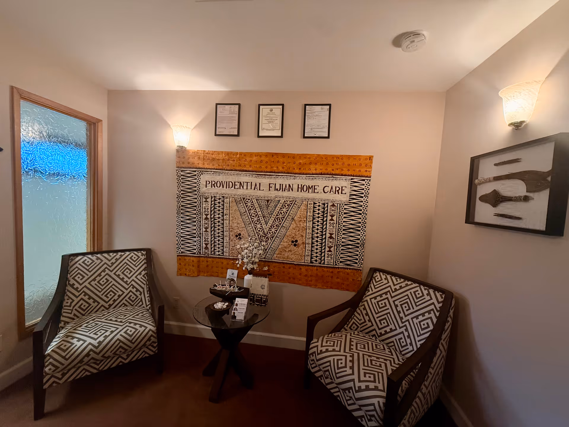 Two patterned armchairs flank a glass table in a waiting area with a wall tapestry.