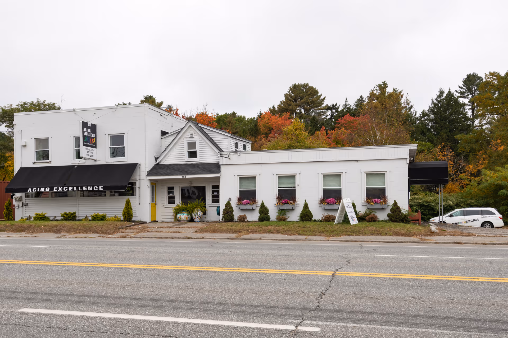 A white, single-story aging care facility with a black awning labeled Aging Excellence along a street.