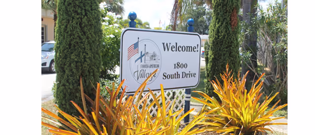Outdoor welcome sign for Finnish-American Village framed by tall shrubs and colorful plants.