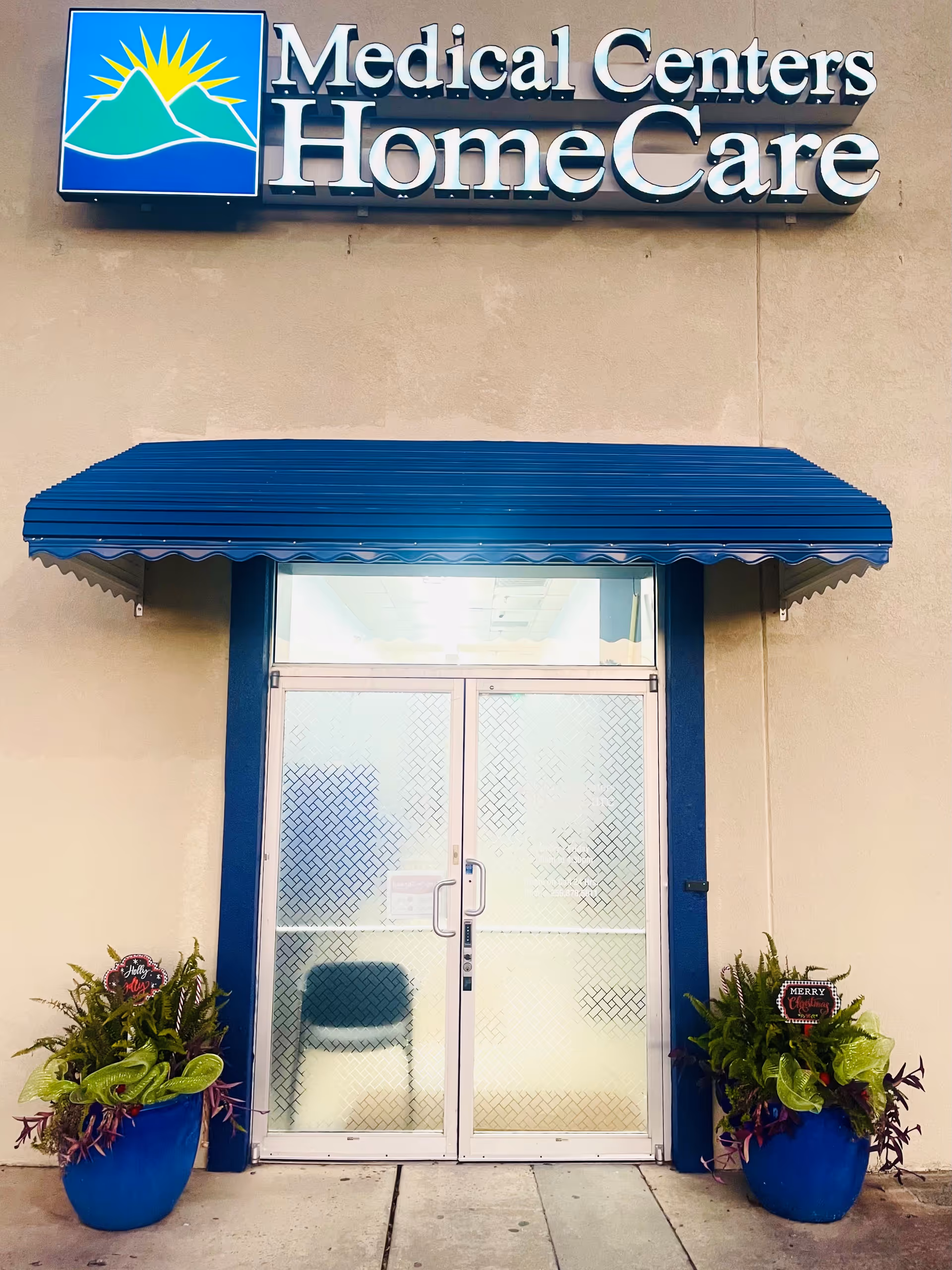 Exterior entrance of Medical Centers HomeCare with glass doors, blue awning, and potted plants.