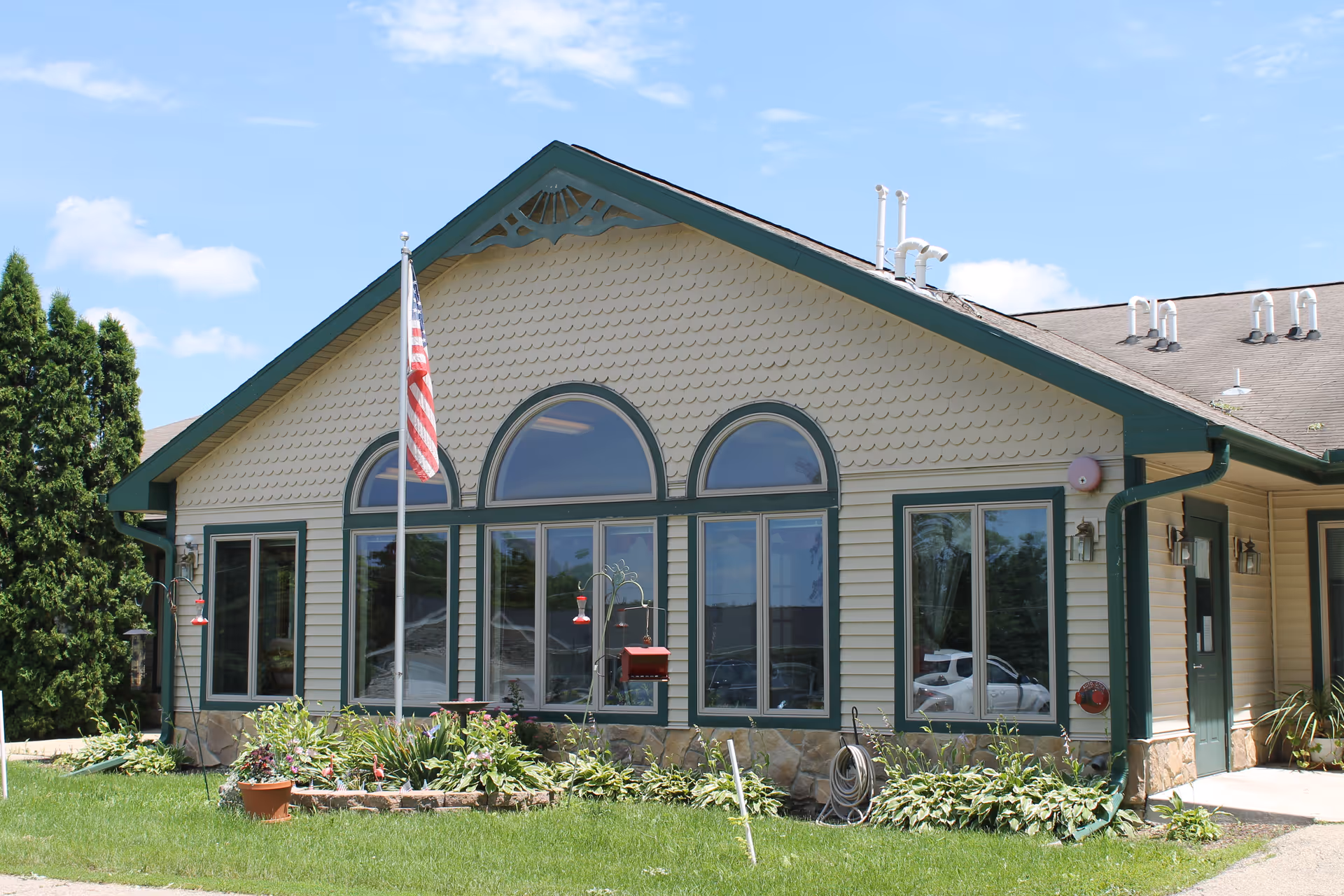 Exterior view of a single-story building with beige siding and green trim, featuring three large arched windows and two rectangular windows. An American flag is mounted on a pole in front of the building, surrounded by a small garden with plants and flowers. The sky is clear with a few clouds.
