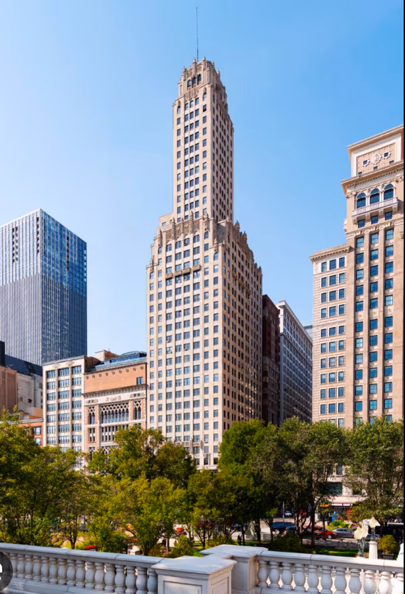 Urban cityscape featuring a tall beige skyscraper, adjacent buildings, and a white railing in foreground.