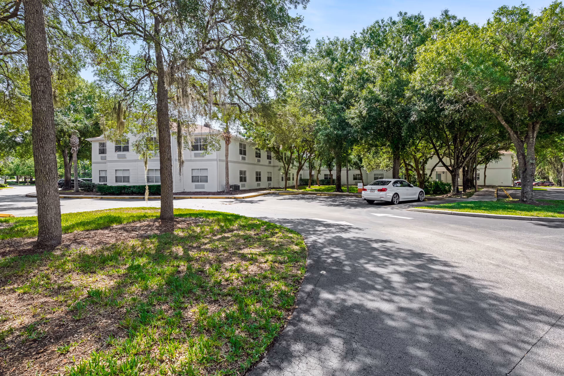 Exterior view of a senior living facility with white two-story buildings surrounded by large trees and greenery. A white car is parked on the paved driveway, and the area is shaded by the trees.