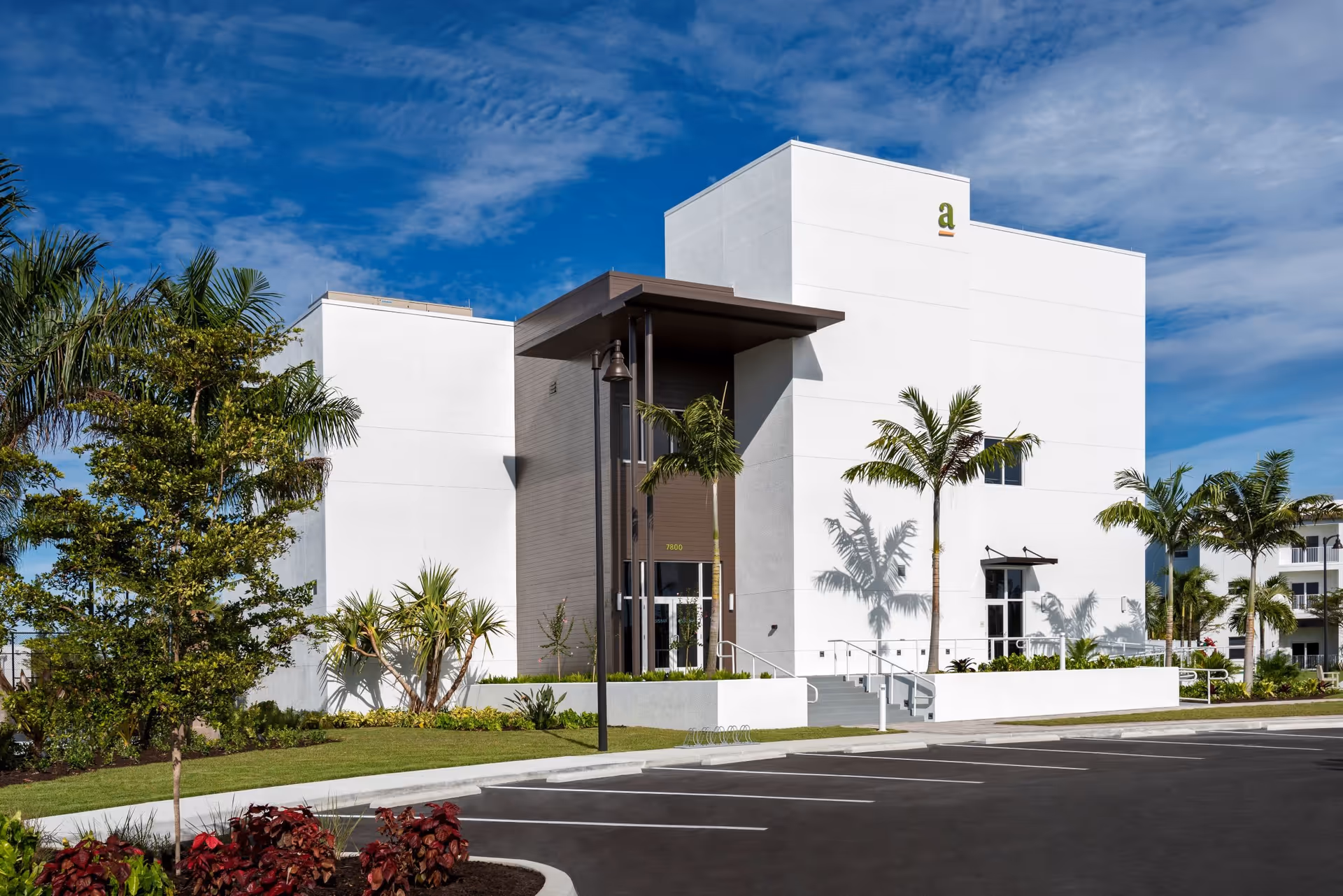Modern white multi-story senior living facility building entrance with palm trees and a parking lot under a blue sky.