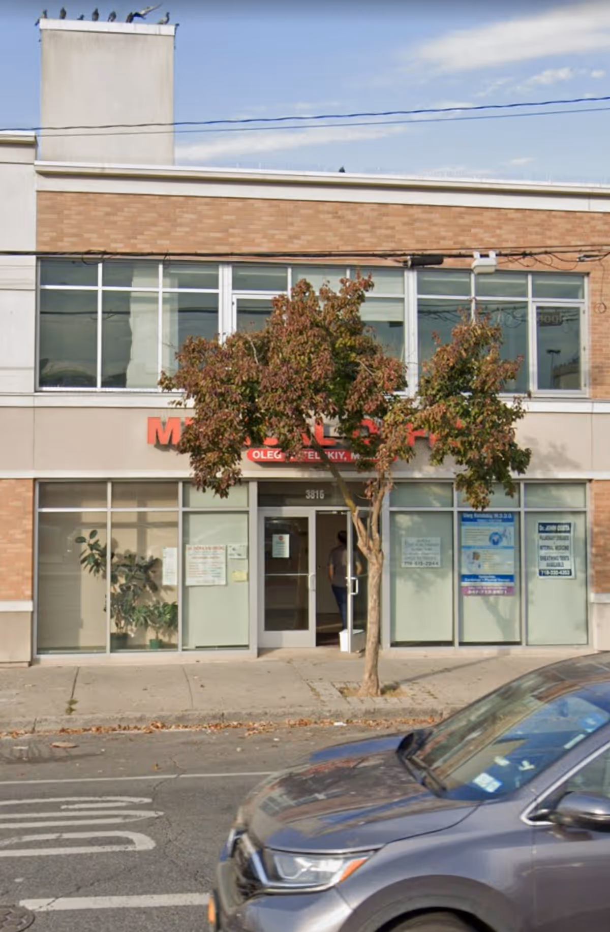 Street-level view of a storefront facility with large windows, a tree, and a car in foreground.