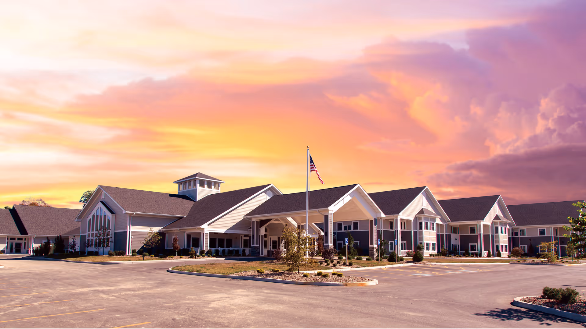 Exterior front view of The Inn at Poland Way senior living building with an American flag and parking lot at sunset.