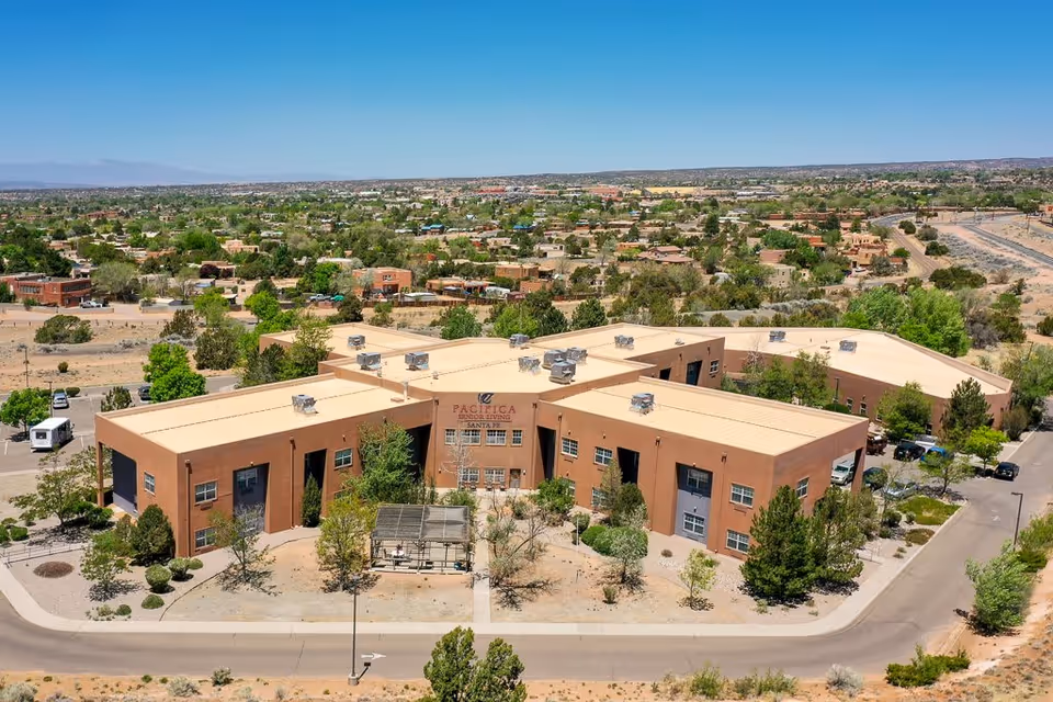 Aerial view of a large, single-story senior living facility with a tan flat roof and brown exterior walls, surrounded by trees and landscaping. The building is situated in a suburban area with many houses and greenery visible in the background under a clear blue sky.