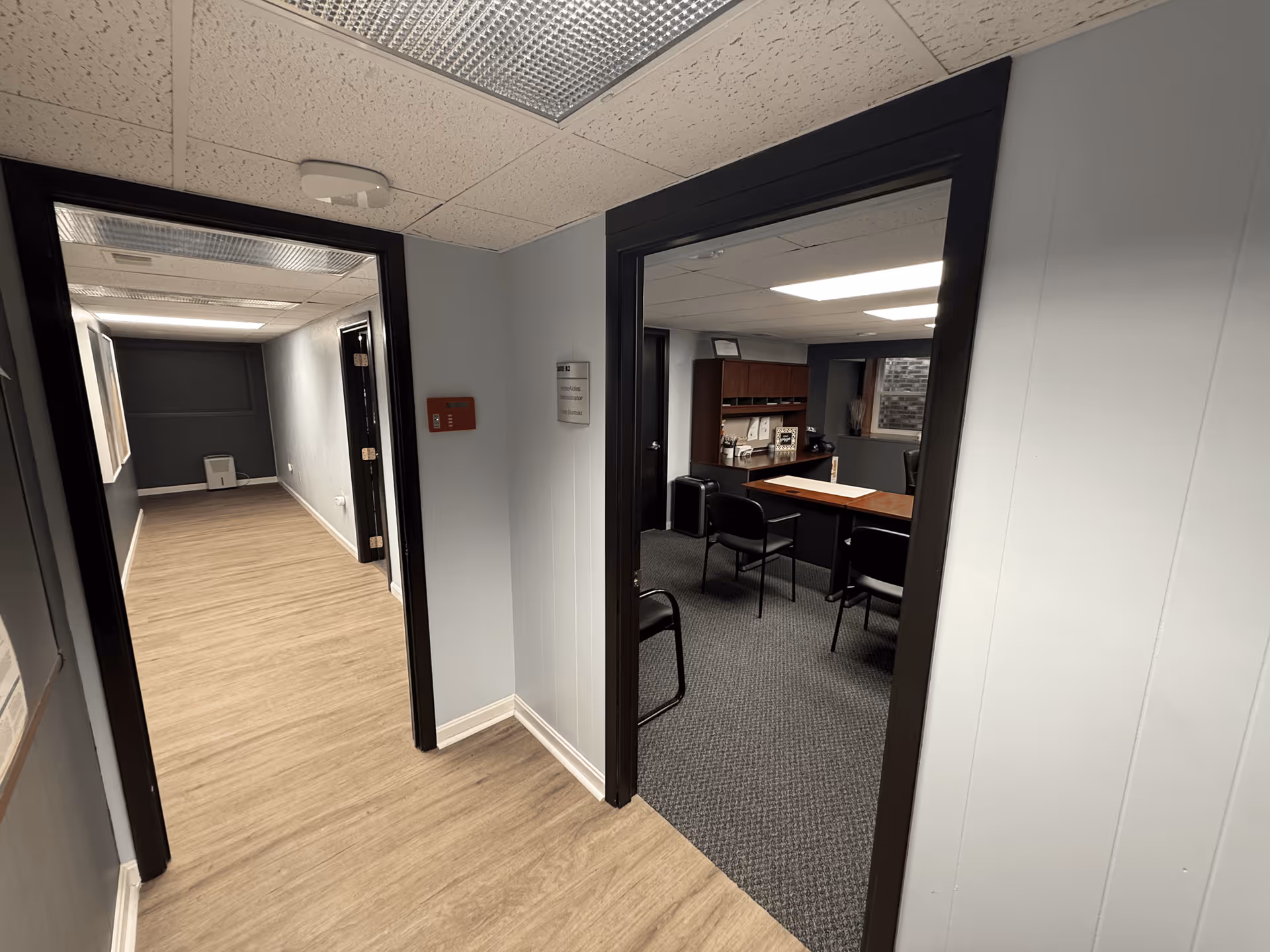 Hallway with wood-like flooring leads to a meeting room containing a desk and chairs.