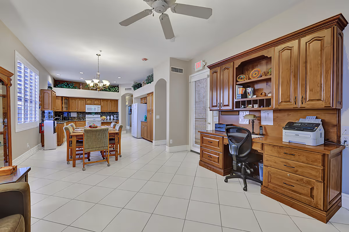 Bright, spacious senior living common area with dining table, kitchen, and wooden desk.