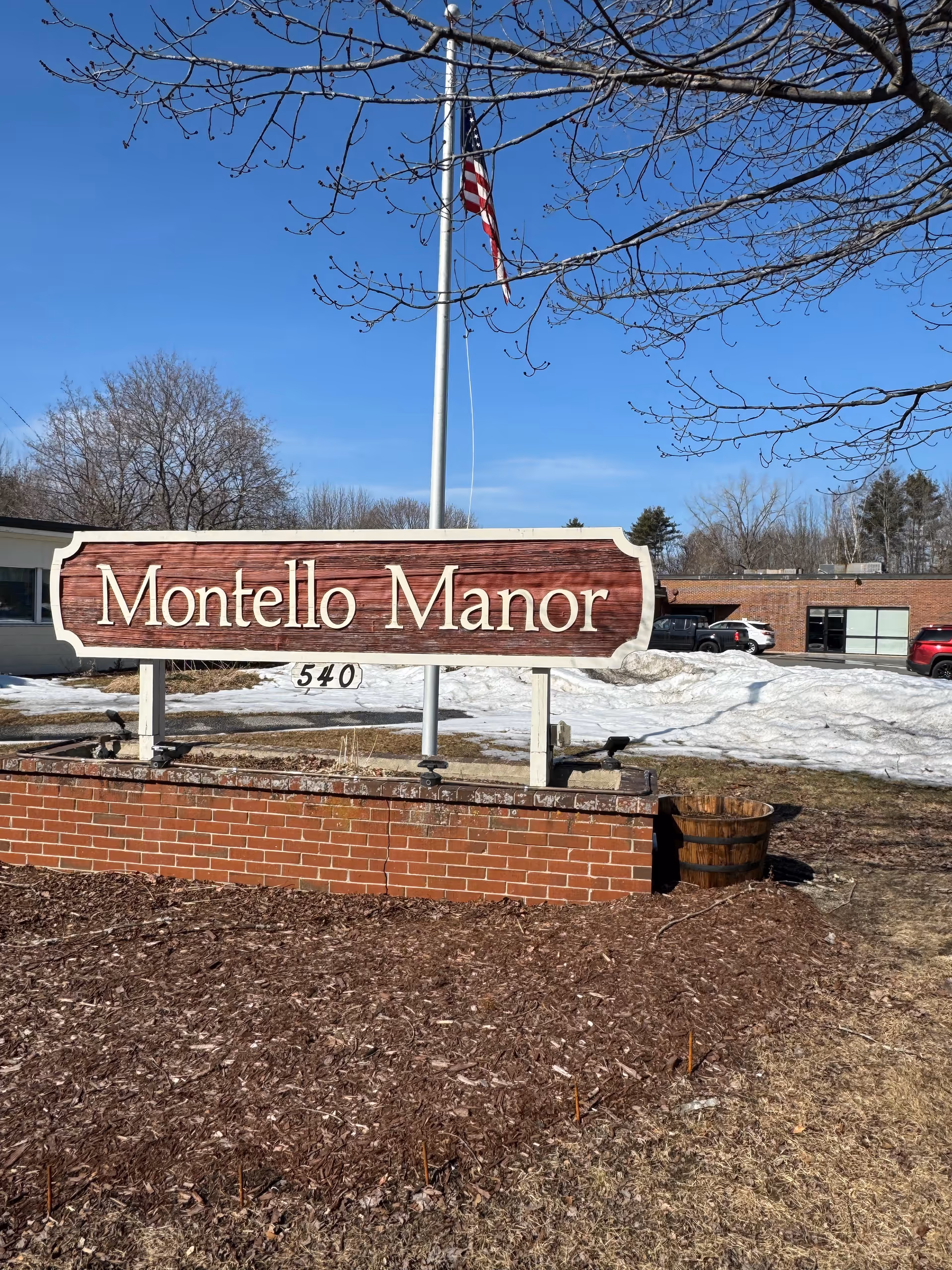 Outdoor view of Montello Manor sign with a flagpole and snowy landscaping.