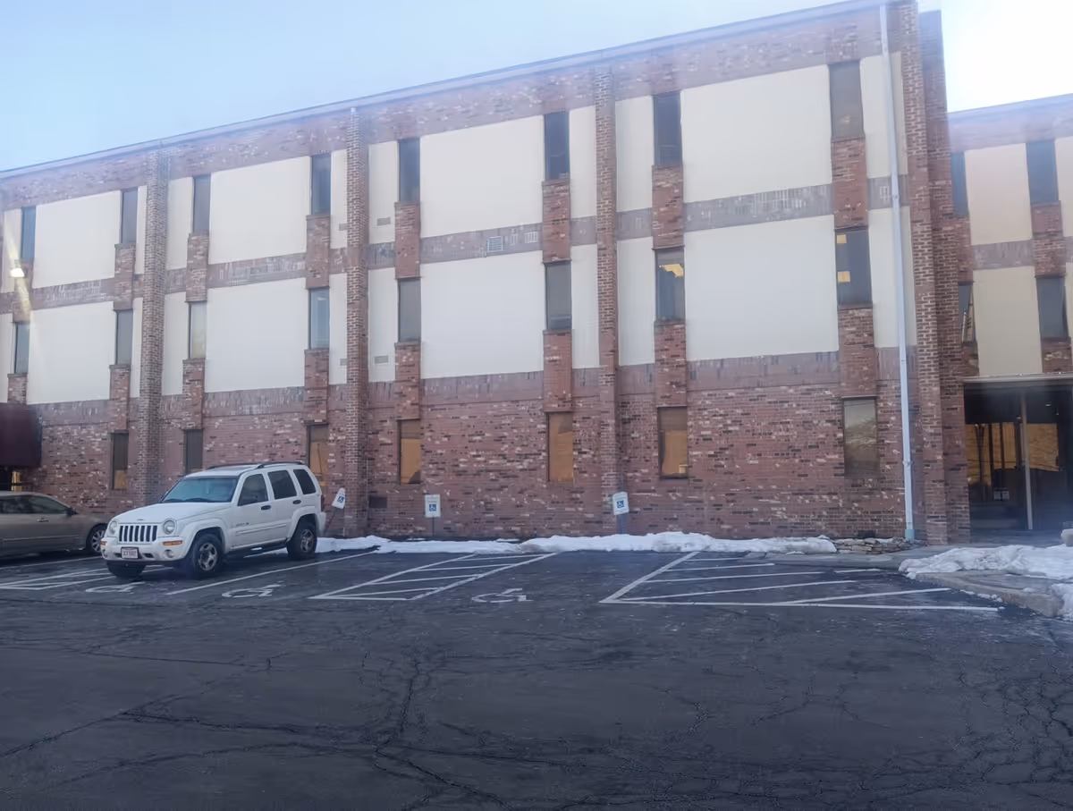Exterior brick facility with boarded windows and a white SUV in the parking lot